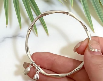 May include: A silver bangle with a flowing, twisted design. Two small charms dangle from the bracelet. The background features green leaves and a white marble surface.