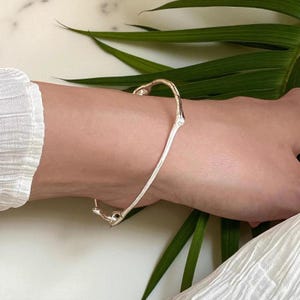 Puede incluir: Una pulsera plateada con un diseño orgánico único. La pulsera se curva alrededor de la muñeca, con una estructura similar a una rama y un pequeño detalle redondeado en el extremo. La pulsera se lleva en un brazo de piel clara, con una manga blanca visible.