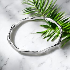 Puede incluir: Un brazalete plateado ondulado. El brazalete tiene un diseño orgánico y fluido. La joya está colocada sobre una superficie de mármol blanco con una fronda de palma verde en el fondo. El diseño del brazalete es suave y reflectante.