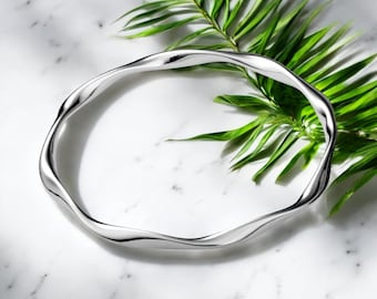 May include: A silver-coloured, wavy bangle bracelet. The bracelet has an organic, fluid design. The jewellery is set against a white marble surface with a green palm frond in the background. The bracelet's design is smooth and reflective.