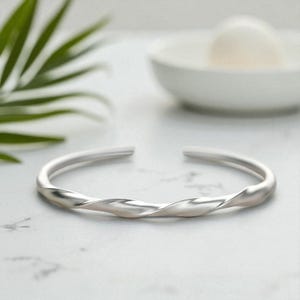 Può includere: Un bracciale a polsino color argento con un design a torsione. Il bracciale aperto è realizzato in metallo liscio e lucido. Il bracciale è su una superficie bianca con uno sfondo sfocato.