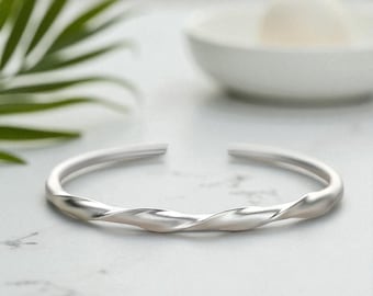 May include: A silver cuff bracelet with a twisted design. The open-ended bracelet is made of a smooth, shiny metal. The bracelet is on a white surface with a blurred background.