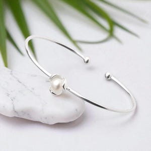 Peut inclure: Un bracelet jonc ouvert argenté avec un motif floral central. La fleur présente une perle blanche en son centre. Le bracelet a un design minimaliste avec des extrémités arrondies. Le fond est une surface semblable à du marbre.