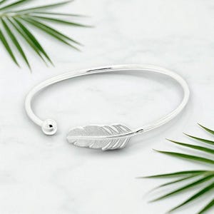 May include: A silver-colored cuff bracelet with a feather design. The bracelet has a smooth, curved band that opens at one end and features a small ball on the other. The feather is detailed with etched lines, adding texture to the design.