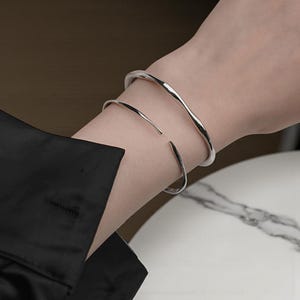Può includere: Due braccialetti color argento sono indossati sul braccio. Il braccialetto superiore ha un design ondulato, mentre il braccialetto inferiore è un semplice bracciale aperto. Il braccio indossa una camicia nera a maniche lunghe.