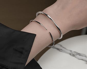 May include: Two silver bracelets are worn on the arm. The top bracelet has a wavy design, while the bottom bracelet is a simple, open cuff. The arm is wearing a black long-sleeve shirt.