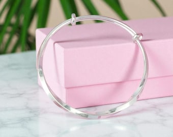 Può includere: Un braccialetto a cerchio argentato con un design a torsione. Il braccialetto è appoggiato su una superficie di marmo bianco e grigio, con una scatola rosa sullo sfondo. Sullo sfondo sono visibili delle foglie verdi.