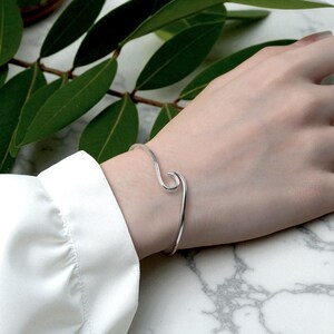 Può includere: Un bracciale a polsino argentato a forma di onda. Il bracciale è indossato su un polso, con una manica di camicia bianca visibile. Le foglie verdi sono sullo sfondo. Il design del bracciale è minimalista ed elegante.