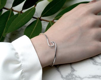 May include: A silver wave-shaped cuff bracelet. The bracelet is worn on a wrist, with a white shirt sleeve visible. Green leaves are in the background. The bracelet's design is minimalist and elegant.