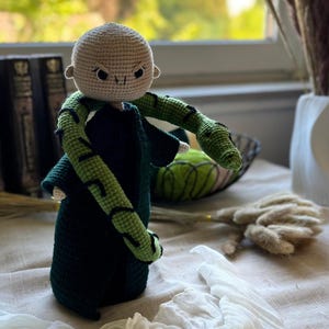Puede incluir: Muñeca de ganchillo hecha a mano con cabeza calva, ojos oscuros y expresión severa. La muñeca lleva una túnica verde oscuro y tiene brazos a rayas verdes y negras. La muñeca está sobre una superficie beige.