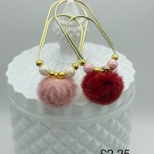 May include: Gold-coloured hoop earrings with a dangling pink and red pom-pom. The earrings feature gold beads and white and pink pearls. The earrings are displayed on a white, textured surface.