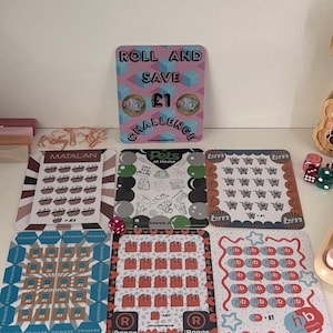 Puede incluir: Una colección de tarjetas de desafío rectangulares y coloridas con varios diseños y texto, incluyendo "Roll and Save £1 Challenge", carritos de compras y logotipos de tiendas. También se ven dados.