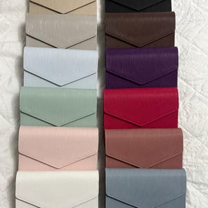 May include: An assortment of envelope wallets in a range of colours: beige, black, brown, purple, red, pink, white, and blue. Each wallet features a textured finish and a flap closure. The wallets are neatly arranged in rows.