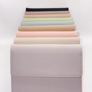 May include: A collection of pastel-colored wallets arranged in a row. The wallets are in shades of lavender, cream, pink, tan, light blue, green, and black. Each wallet has a textured surface and a stitched border, suggesting a leather or leather-like material.