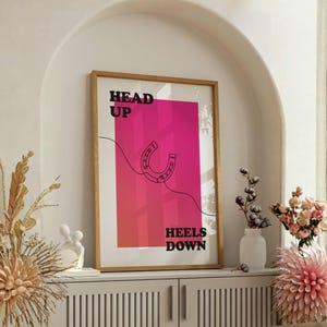 Könnte beinhalten: Ein gerahmter Kunstdruck mit Holzrahmen. Der Druck zeigt einen rosa-roten Farbverlaufshintergrund mit einer Hufeisenillustration und den Worten "HEAD UP" und "HEELS DOWN" in schwarzem Text. Das Kunstwerk wird auf einem weißen Schrank ausgestellt.