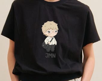 May include: Black crew neck t-shirt featuring a cartoon illustration of a character with glasses, a bag, and the letters "JMN" below the image. The character is wearing a white shirt and black pants. The shirt is short-sleeved.