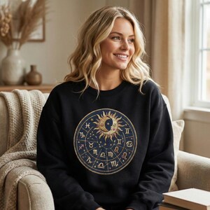 May include: Black sweatshirt featuring a circular zodiac design with a sun, moon, and star graphic. The design is in gold and blue tones. The sweatshirt is made of a soft material.
