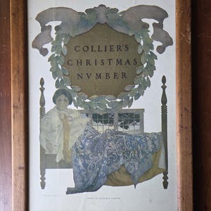 May include: Framed vintage print with a wooden frame. The print features the text "COLLIER'S CHRISTMAS NUMBER" within a decorative wreath. A bed with a blue patterned blanket and a person in a white garment are also depicted.