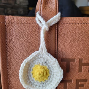 May include: A handmade crochet egg-shaped charm with a white outer edge and a yellow center, hanging from a brown leather bag handle. The charm is attached with a white yarn loop.