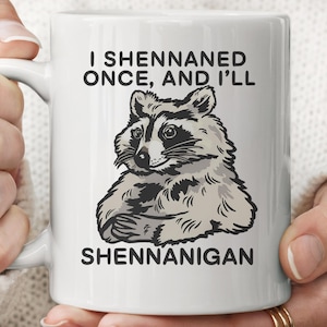 Pode incluir: Caneca de cerâmica branca com ilustração de um guaxinim e o texto "I Shennaned Once, And I'll Shennanigan". O guaxinim é em tons de cinza e branco. Ideal para café ou chá.