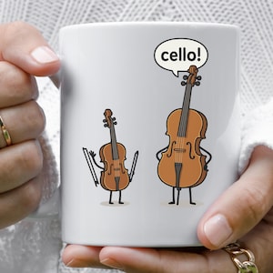 May include: White ceramic mug featuring a cartoon illustration of a violin and a cello. The cello has a speech bubble that says "cello!". The instruments have arms, legs, and facial expressions.