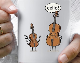 May include: White ceramic mug featuring a cartoon illustration of a violin and a cello. The cello has a speech bubble that says "cello!". The instruments have arms, legs, and facial expressions.