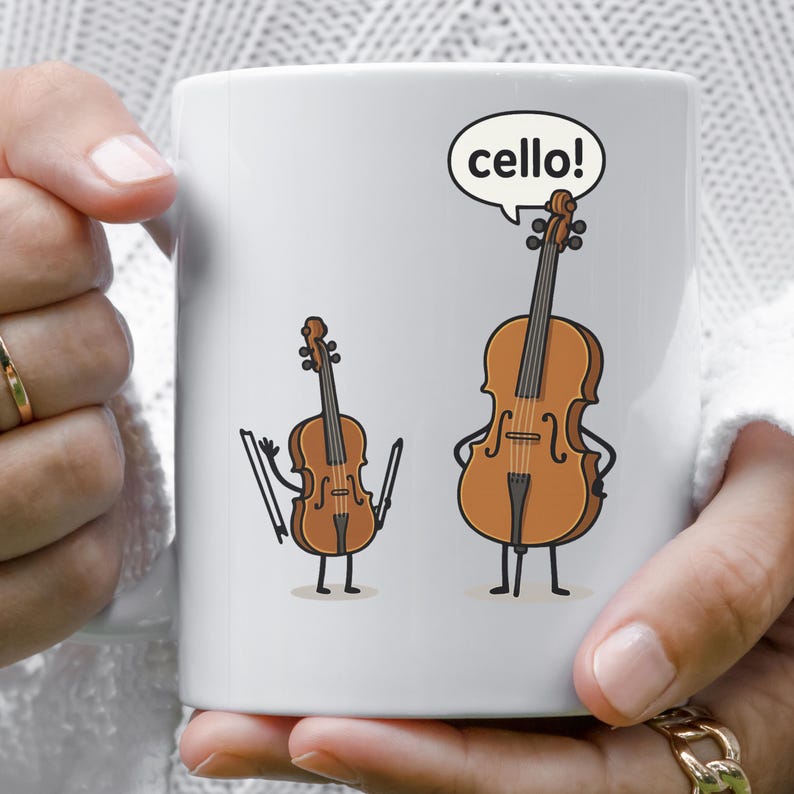 May include: White ceramic mug featuring a cartoon illustration of a violin and a cello. The cello has a speech bubble that says "cello!". The instruments have arms, legs, and facial expressions.