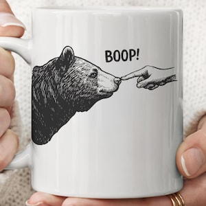 May include: White ceramic mug featuring a black and white illustration of a bear being poked on the nose by a finger. The word "BOOP!" is printed above the finger.