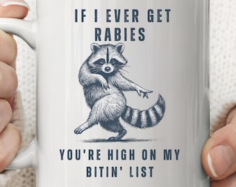May include: White ceramic mug with a black illustration of a raccoon and text that reads "IF I EVER GET RABIES YOU'RE HIGH ON MY BITIN' LIST". The raccoon is depicted in a dancing pose.