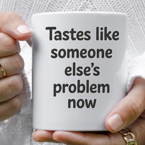 以下が含まれることがあります： 「Tastes like someone else's problem now」の黒い文字が入った白いセラミックマグ。白いセーターと金のジュエリーを身につけた人がマグカップを持っています。