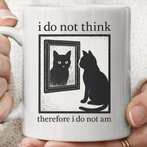 May include: White ceramic mug featuring a black cat looking at its reflection in a mirror. The mug has the text "i do not think" above the image and "therefore i do not am" below. The design is a humorous take on philosophy.