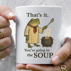May include: White ceramic mug featuring a cartoon frog chef holding a spoon over a steaming pot of soup. The mug has the text "That's it. You're going in the SOUP."