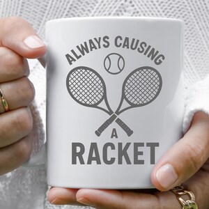May include: White ceramic mug with a tennis theme. The mug features a tennis ball and two crossed tennis rackets in gray, with the words "ALWAYS CAUSING A RACKET" printed in gray.