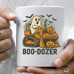 Pode incluir: Caneca de cerâmica branca com um design temático de Halloween. O design inclui um fantasma de desenho animado dirigindo uma escavadeira laranja, com abóboras na caçamba dianteira e morcegos voando acima. O texto "BOO-DOZER" é impresso abaixo.