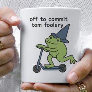 May include: White ceramic mug featuring a green frog riding a scooter and wearing a blue wizard hat. The mug has the text "off to commit tom foolery" in black lettering.