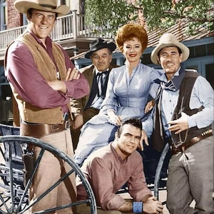 May include: A group of six people, dressed in Western attire, pose for a photo. The men wear cowboy hats, vests, and boots. The woman wears a light blue dress. The background includes a building and a wagon wheel. The image is in color.