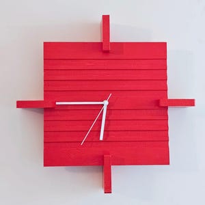 May include: A square, red wooden wall clock with white hands. The clock has a minimalist design with horizontal wooden slats and four rectangular extensions. The clock is mounted on a white wall.