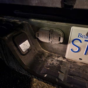 May include: Close-up of a vehicle's rear bumper, featuring a trailer hitch connector and a license plate. The license plate reads "Beautiful ST" with blue lettering. The bumper is dark gray and shows signs of wear.