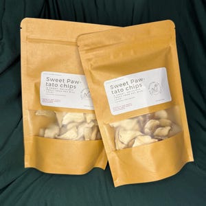 May include: Two brown paper bags of "Sweet Paw-tato Chips" for pets. Each bag has a clear window showing the sweet potato chips inside. The bags are sealed and have a white label with text and a logo. The background is a dark green fabric.