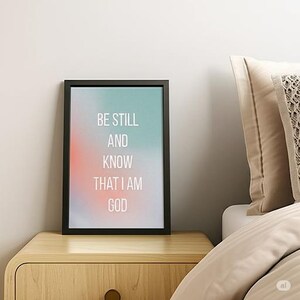 May include: A framed print with the words "BE STILL AND KNOW THAT I AM GOD" in white lettering. The background has a gradient of colors, including teal, coral, and white. The frame is black, and the print is displayed on a wooden nightstand.