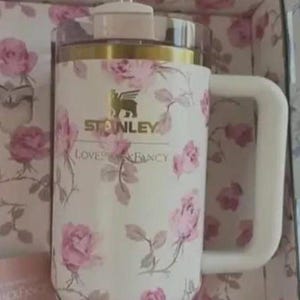 May include: A white Stanley travel mug with a floral rose pattern and a gold rim. The mug has a white handle and a pink bow on the lid. The text "Stanley Loves Fancy" is printed on the mug.