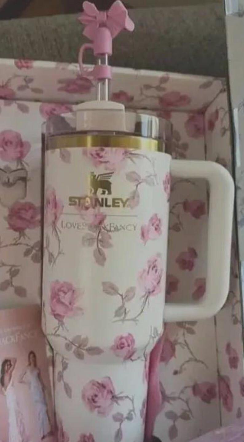 May include: A white Stanley travel mug with a floral rose pattern and a gold rim. The mug has a white handle and a pink bow on the lid. The text "Stanley Loves Fancy" is printed on the mug.