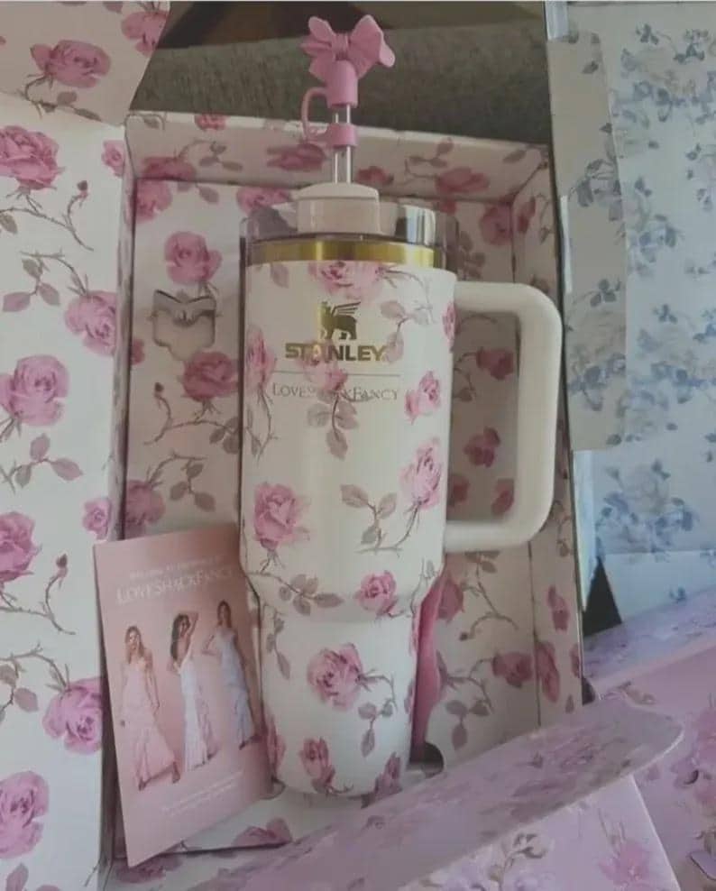 May include: A white Stanley tumbler with a pink rose pattern, a white handle, and a pink bow-shaped topper. The tumbler is in a matching floral-printed box with the text "Stanley Loves Shack Fancy". A card with images of women in dresses is also visible.