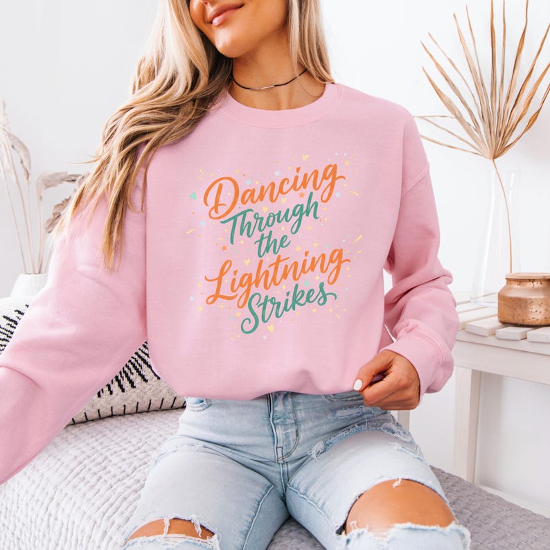 May include: A light pink sweatshirt with the text "Dancing Through the Lightning Strikes" in orange, green, and blue. The sweatshirt has long sleeves and a crew neck. The text is in a script font.