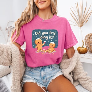 Puede incluir: Una camiseta magenta con el texto "Did you try icing it?" y un diseño de hombre de jengibre de dibujos animados. Un hombre de jengibre está de pie y sosteniendo glaseado, mientras que el otro está acostado. La camiseta se combina con pantalones cortos de mezclilla.