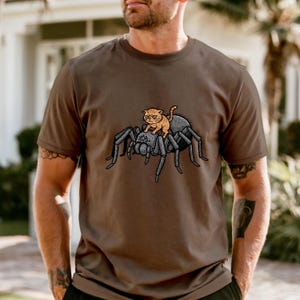 May include: Brown t-shirt featuring a cartoon illustration of an orange cat riding a large gray spider. The cat has a grumpy expression. The spider has multiple legs and a detailed design.