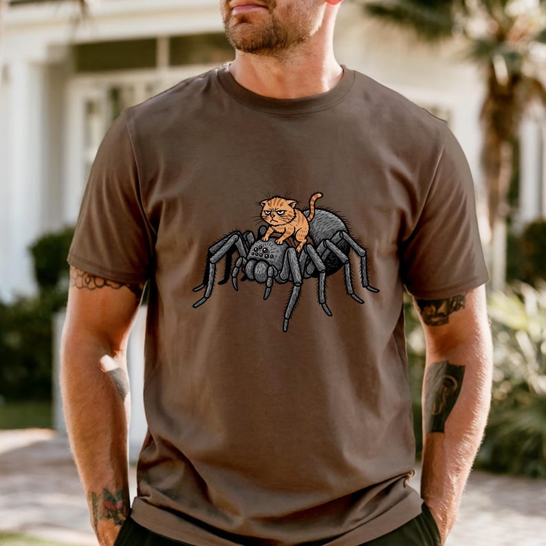 May include: Brown t-shirt featuring a cartoon illustration of an orange cat riding a large gray spider. The cat has a grumpy expression. The spider has multiple legs and a detailed design.