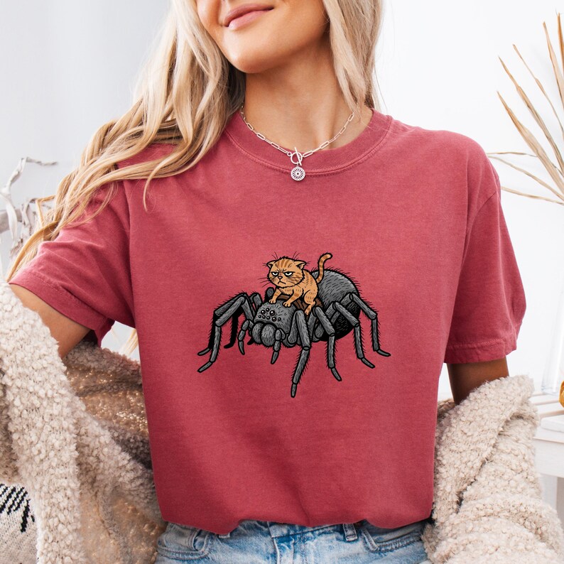May include: A maroon t-shirt featuring a graphic of a black spider with an orange cat riding on its back. The cat has an angry expression. The shirt has short sleeves and a crew neck.