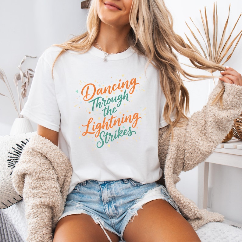 May include: White t-shirt with the text "Dancing Through the Lightning Strikes" in orange and teal script. The shirt has a crew neck and short sleeves. The design includes colorful starburst accents.