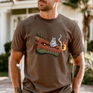 May include: Brown t-shirt featuring a cartoon dragon with red, orange, and green scales, and a gray cat sitting on its snout. The dragon is breathing fire. The shirt is a casual wear item.